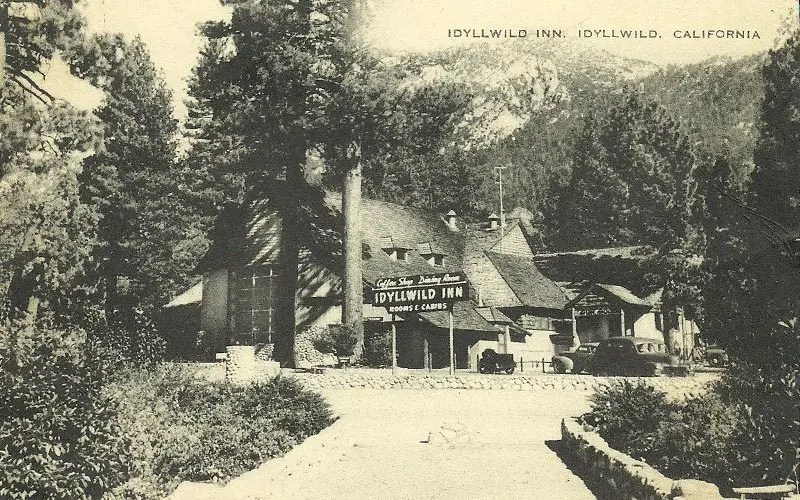 A black and white photograph of the original Idyllwild Inn among the pines, its sign reading Idyllwild Inn, Rooms and Cabins, with early motor cars parked outside