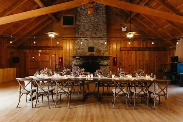 A long banquet table set with flowers and cross-back chairs before the tall stone fireplace in the wood-panelled hall at Tahquitz Pines Retreat