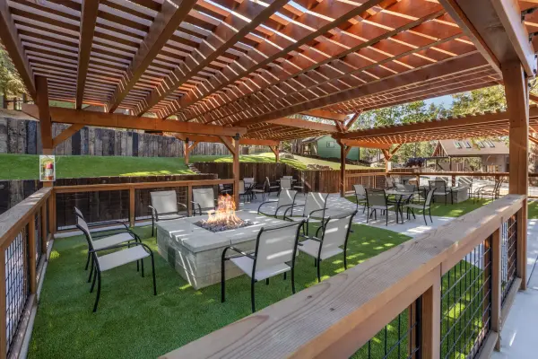 Lounge chairs around a lit fire table under a timber pergola at Tahquitz Pines Retreat, a green lawn and pines beyond the railing