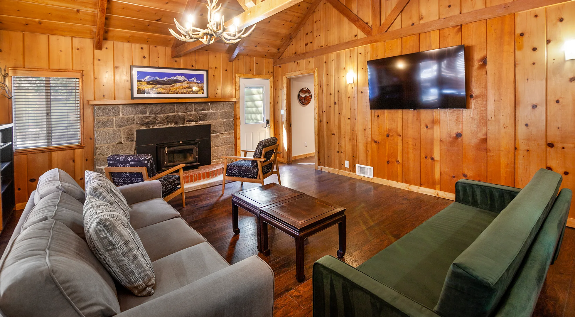 A cabin great room at Tahquitz Pines Retreat with an antler chandelier, a stone fireplace, a wall-mounted television and two sofas under a vaulted pine ceiling