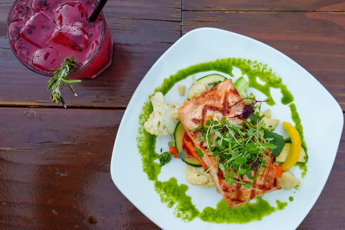 A plate of grilled salmon on vegetables with a herb sauce, beside a berry drink on a dark wood table at The Peak Restaurant