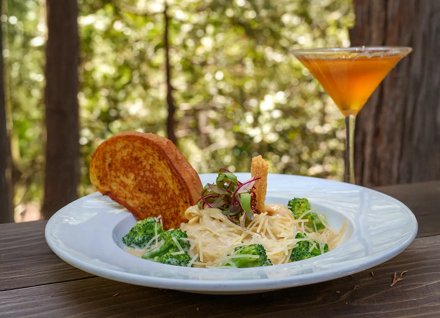 A bowl of pasta with broccoli and garlic toast beside an amber cocktail on a wooden patio table under the pines at The Peak Restaurant