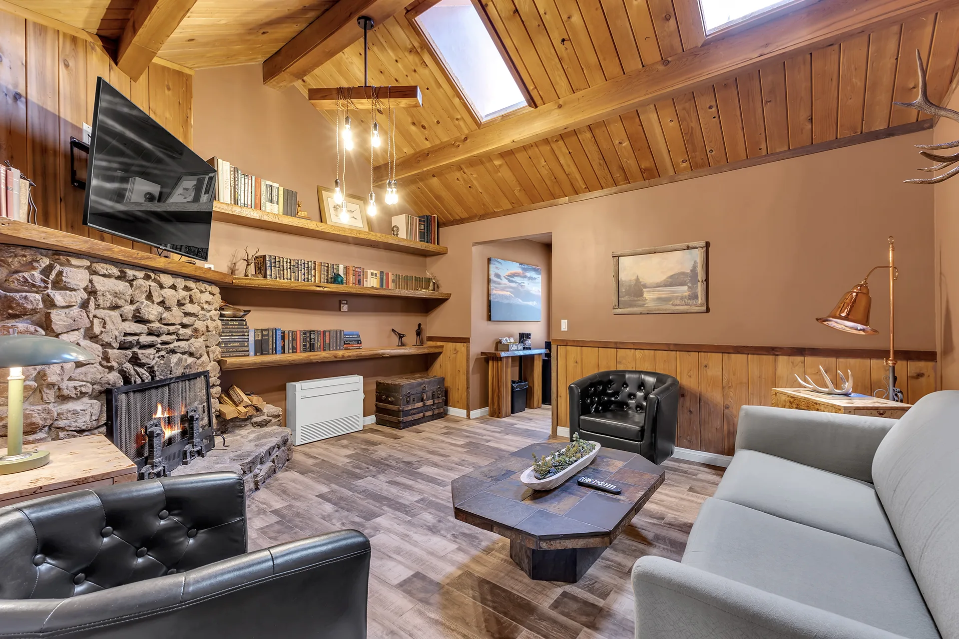 The lounge at Strawberry Creek Inn, a stone fireplace lit under a wood-panelled ceiling with skylights, shelves of books and leather armchairs