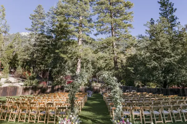 Rows of wooden chairs set for a wedding ceremony on the lawn at Tahquitz Pines Retreat, a flower-covered arch at the aisle and pines and granite behind