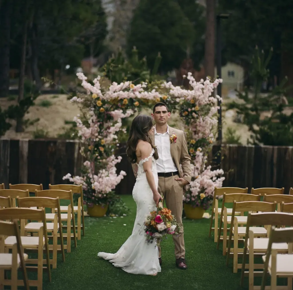 A bride and groom beneath a blossom and rose ceremony arch on the lawn at Tahquitz Pines Retreat, rows of wooden chairs either side