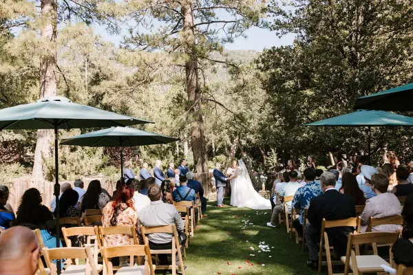 Guests seated under green umbrellas watch a wedding ceremony on the lawn at Tahquitz Pines Retreat, tall pines and the mountainside behind