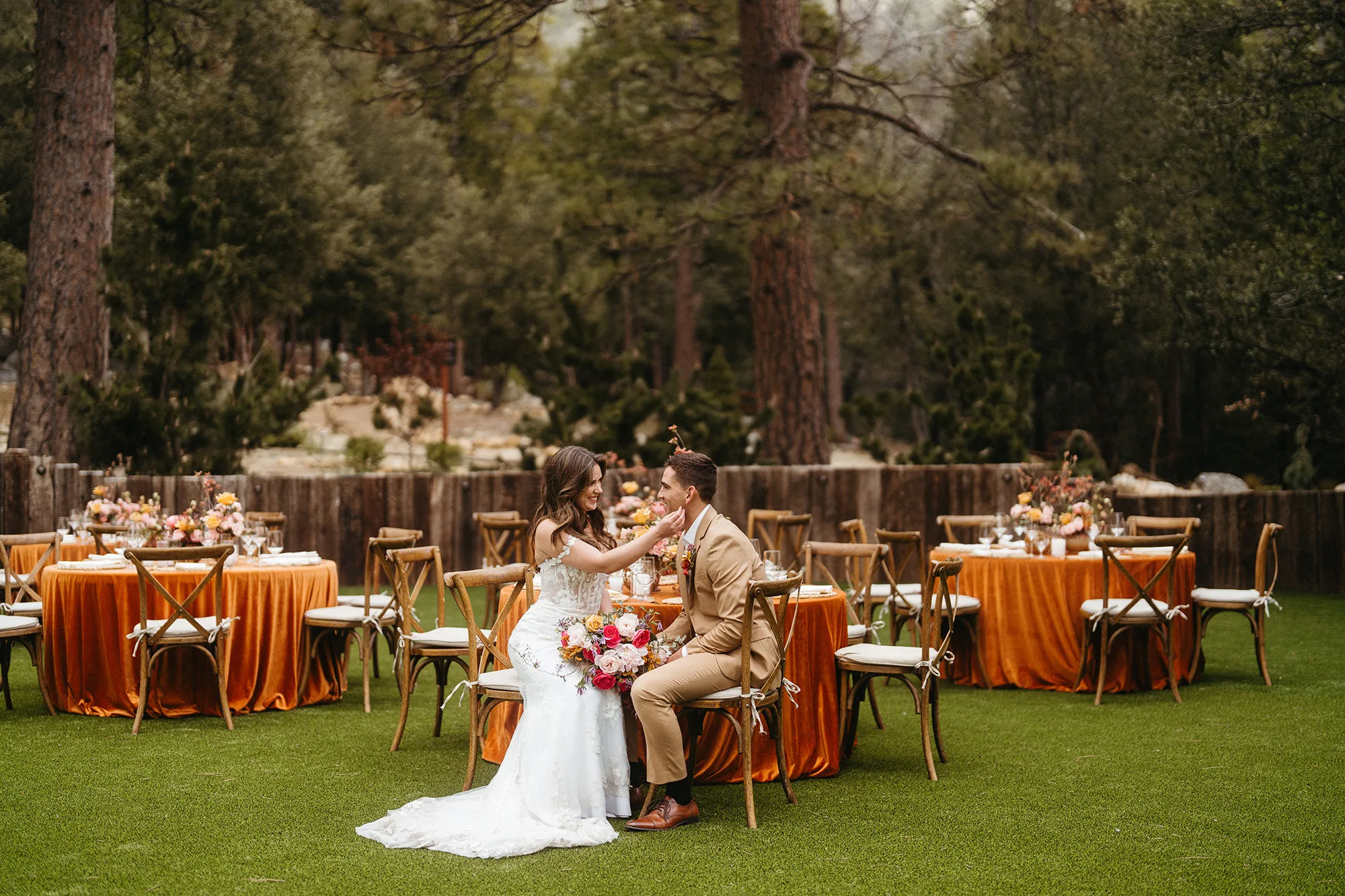 A bride and groom sitting together at their reception on the lawn, orange-linened tables and tall pines around them